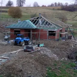 Wznoszony budynek z czerwonej cegły z częściowo ukończonym dachem, widoczny niebieski pojazd terenowy i sprzęt budowlany na nierównym terenie w pochmurny dzień.