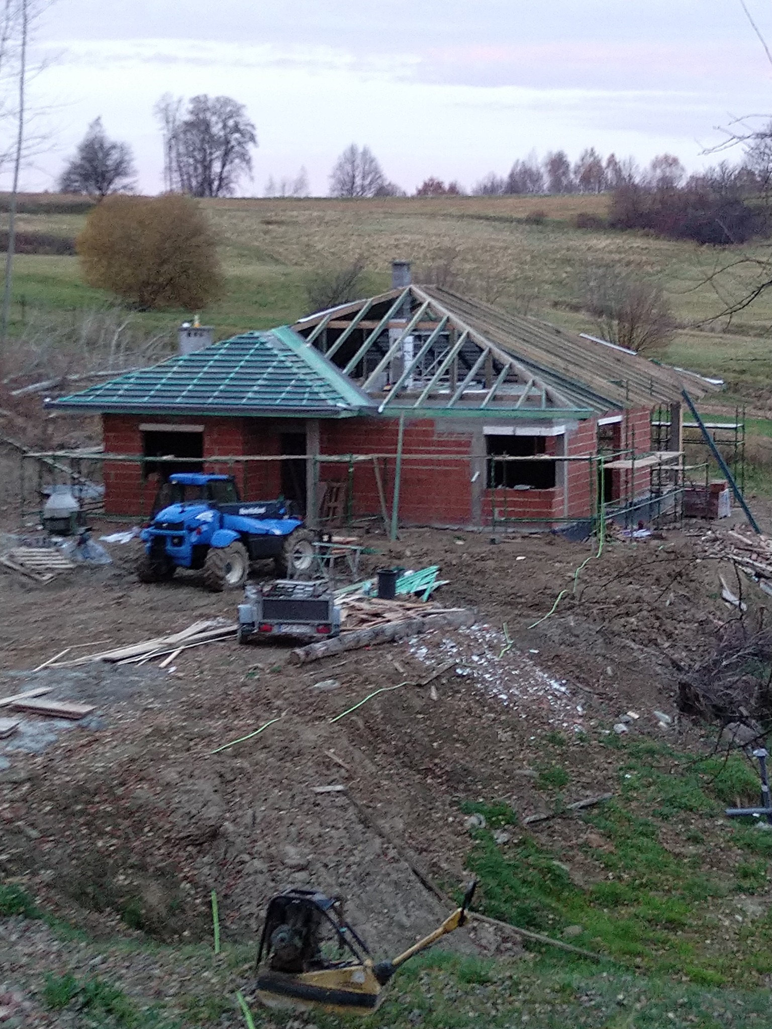 Wznoszony budynek z czerwonej cegły z częściowo ukończonym dachem, widoczny niebieski pojazd terenowy i sprzęt budowlany na nierównym terenie w pochmurny dzień.