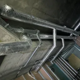 Widok z dołu na izolowane, srebrne rury wentylacyjne pod betonowym sufitem w trakcie montażu, widoczne metalowe korytka kablowe i elementy konstrukcyjne sufitu podwieszanego.