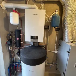 Biały piec gazowy Vaillant z podłączonymi rurami miedzianymi i czarnym zbiornikiem buforowym, widoczne także białe i srebrne rury wentylacyjne oraz niebieski zbiornik wyrównawczy, na podłodze leży...