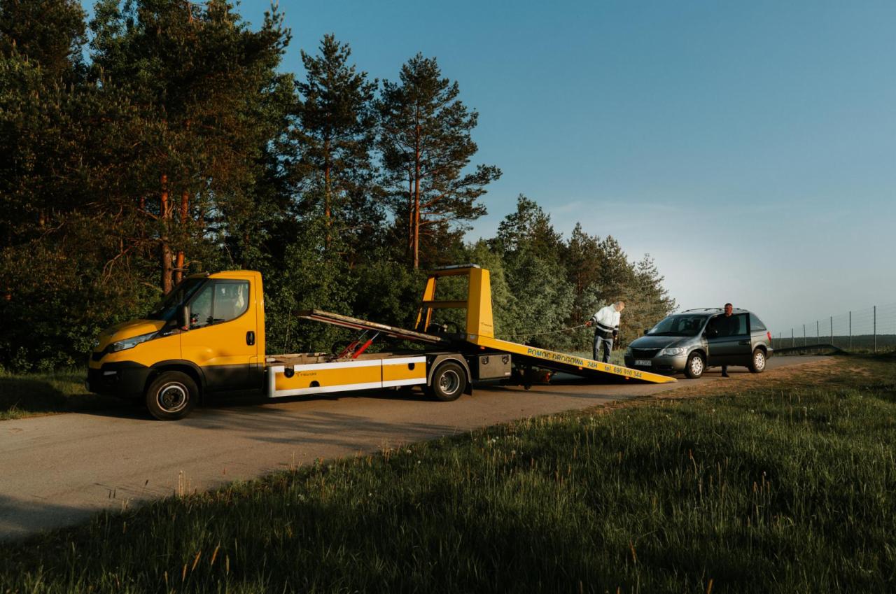 Żółta laweta z naczepą holuje szarego minivana na poboczu drogi w słoneczny dzień. Widoczny mężczyzna w białej kurtce i otwarte drzwi minivana.