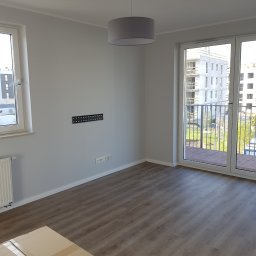 Reno-Art Rafał Łużyński - Pomalowane na szaro wnętrze pokoju we Wrocławiu z panelami na podłodze i przymocowanym uchwytem na telewizor na ścianie, widok na balkon i budynek w budowie za oknem.