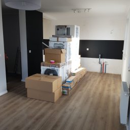 Reno-Art Rafał Łużyński - Puste pomieszczenie z nową podłogą, stosem zapakowanych urządzeń AGD i RTV, oraz fragmentem ściany pomalowanej na czarno, przygotowane do dalszych prac wykończeniowych.