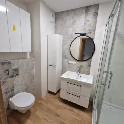 Nowoczesna łazienka z drewnopodobną podłogą, wiszącą toaletą, szafką z umywalką, wysoką szafką i kabiną prysznicową ze szklanymi drzwiami, okrągłe lustro z czarną ramą.