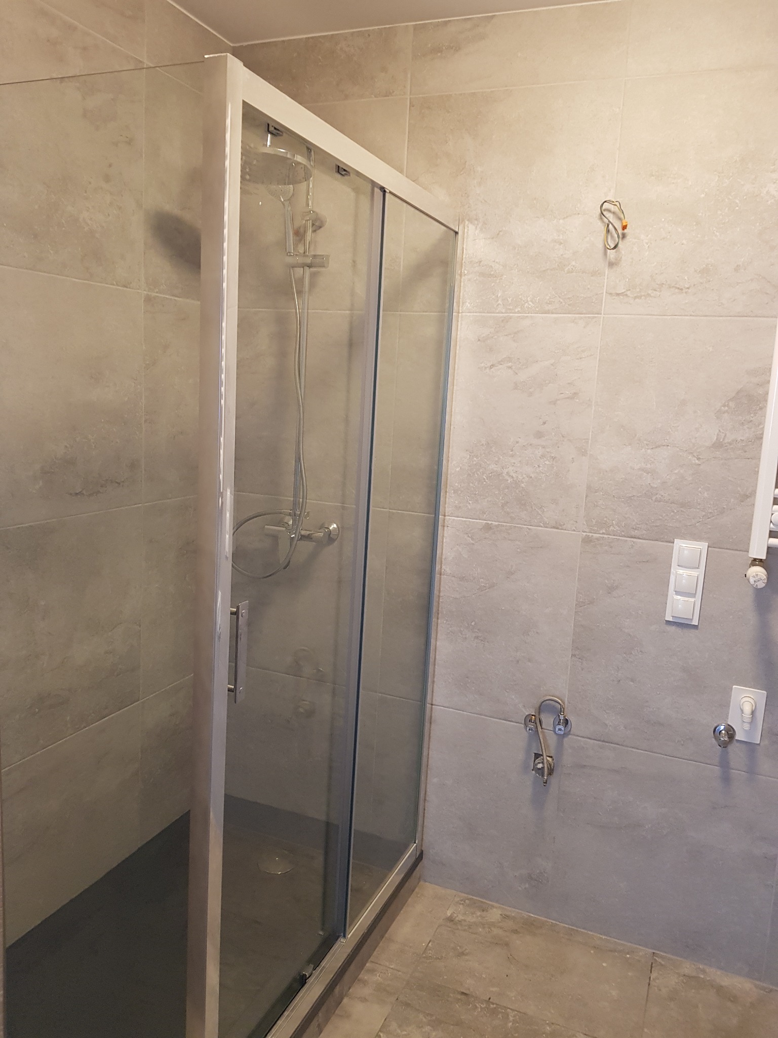 Nowoczesna kabina prysznicowa z deszczownicą, obudowana jasnymi, kwadratowymi płytkami imitującymi kamień, z widocznymi przyłączami wodnymi i elementami instalacji elektrycznej na ścianie.