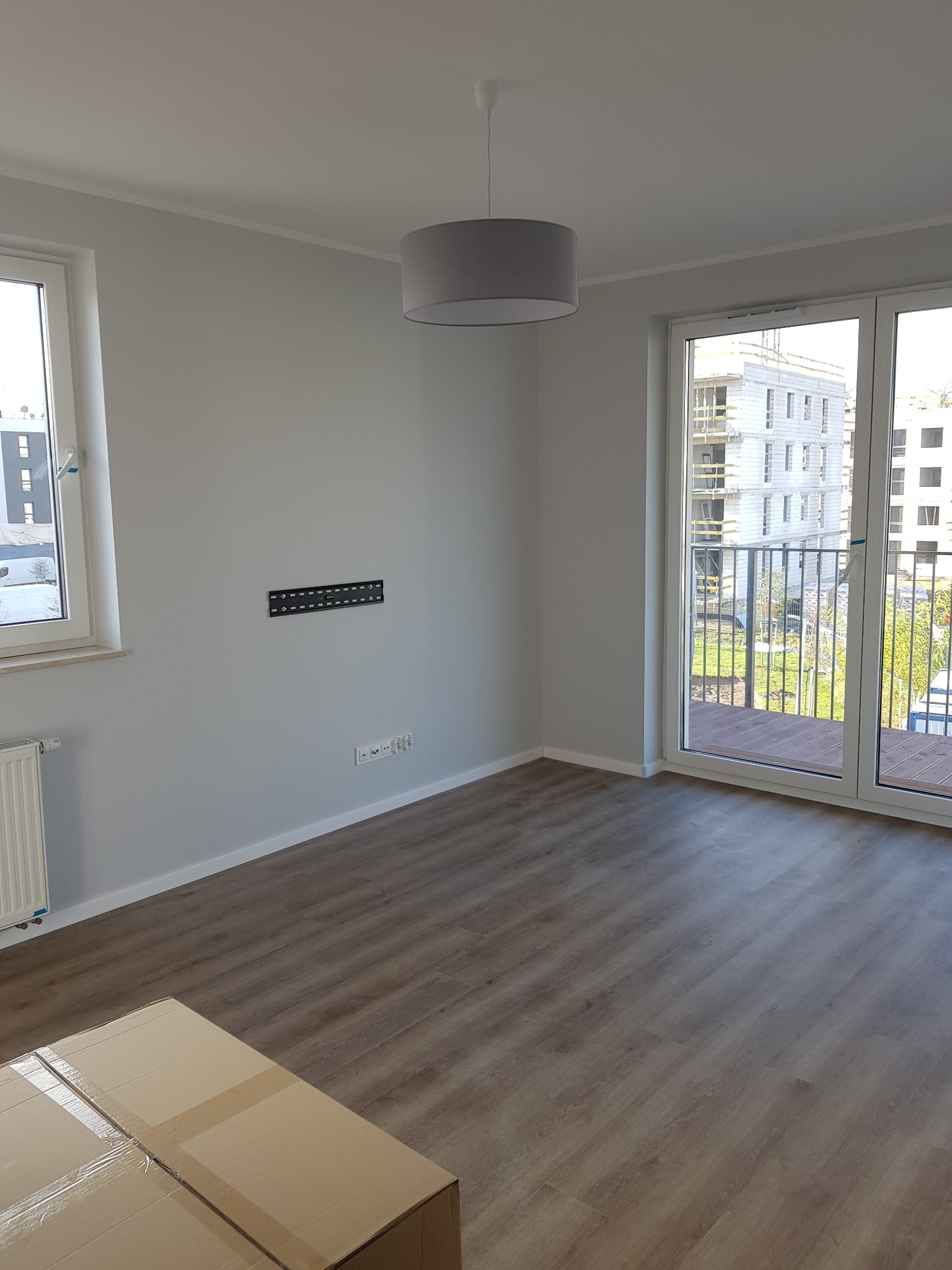 Pomalowane na szaro wnętrze pokoju we Wrocławiu z panelami na podłodze i przymocowanym uchwytem na telewizor na ścianie, widok na balkon i budynek w budowie za oknem.
