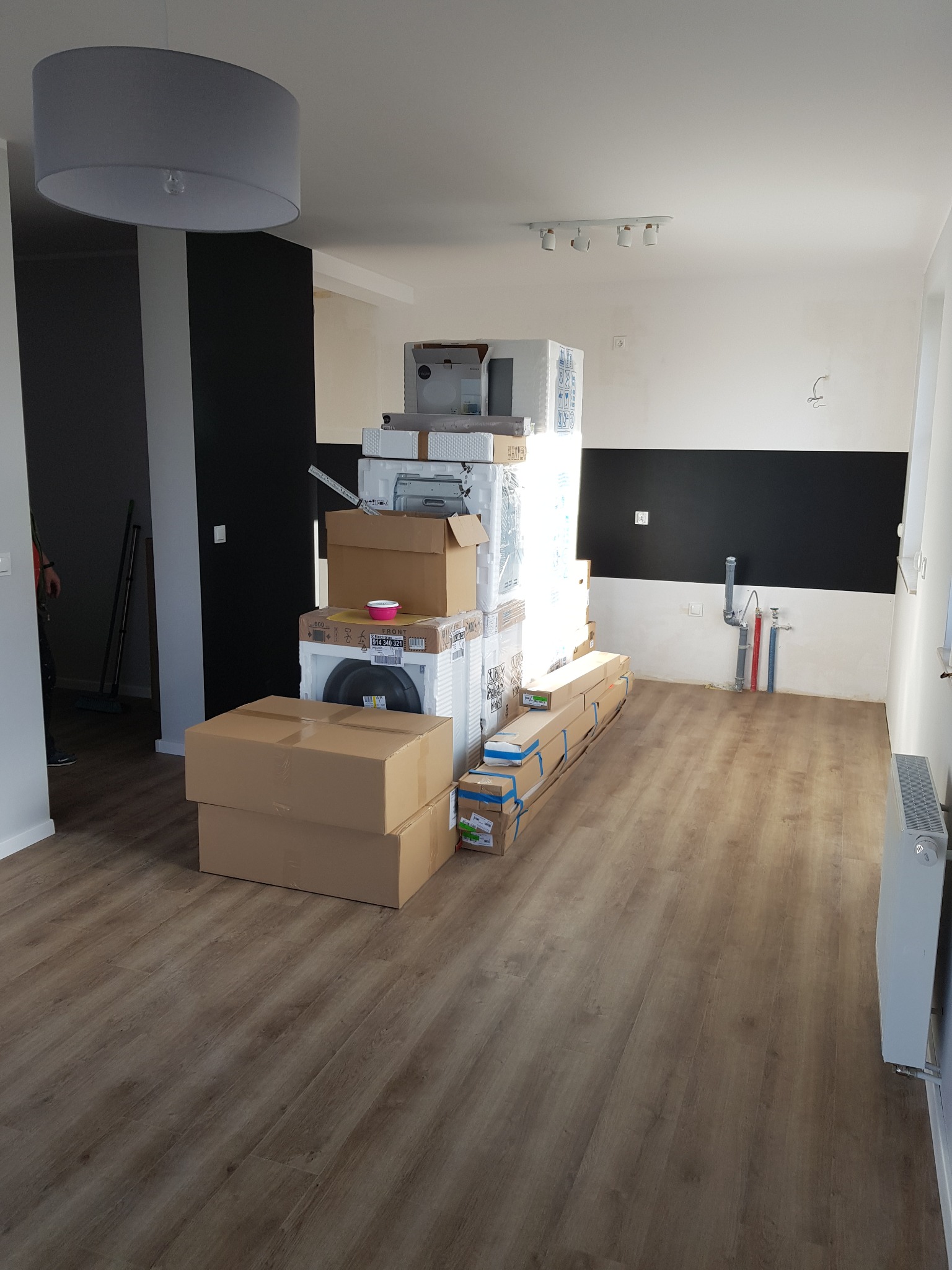 Puste pomieszczenie z nową podłogą, stosem zapakowanych urządzeń AGD i RTV, oraz fragmentem ściany pomalowanej na czarno, przygotowane do dalszych prac wykończeniowych.