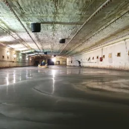 Wnętrze hali z wylewaną, mokrą posadzką; dw&oacute;ch pracownik&oacute;w w oddali używa maszyn do wyr&oacute;wnywania powierzchni, widoczne odbicia światła w mokrej posadzce, ściany hali z oknami.