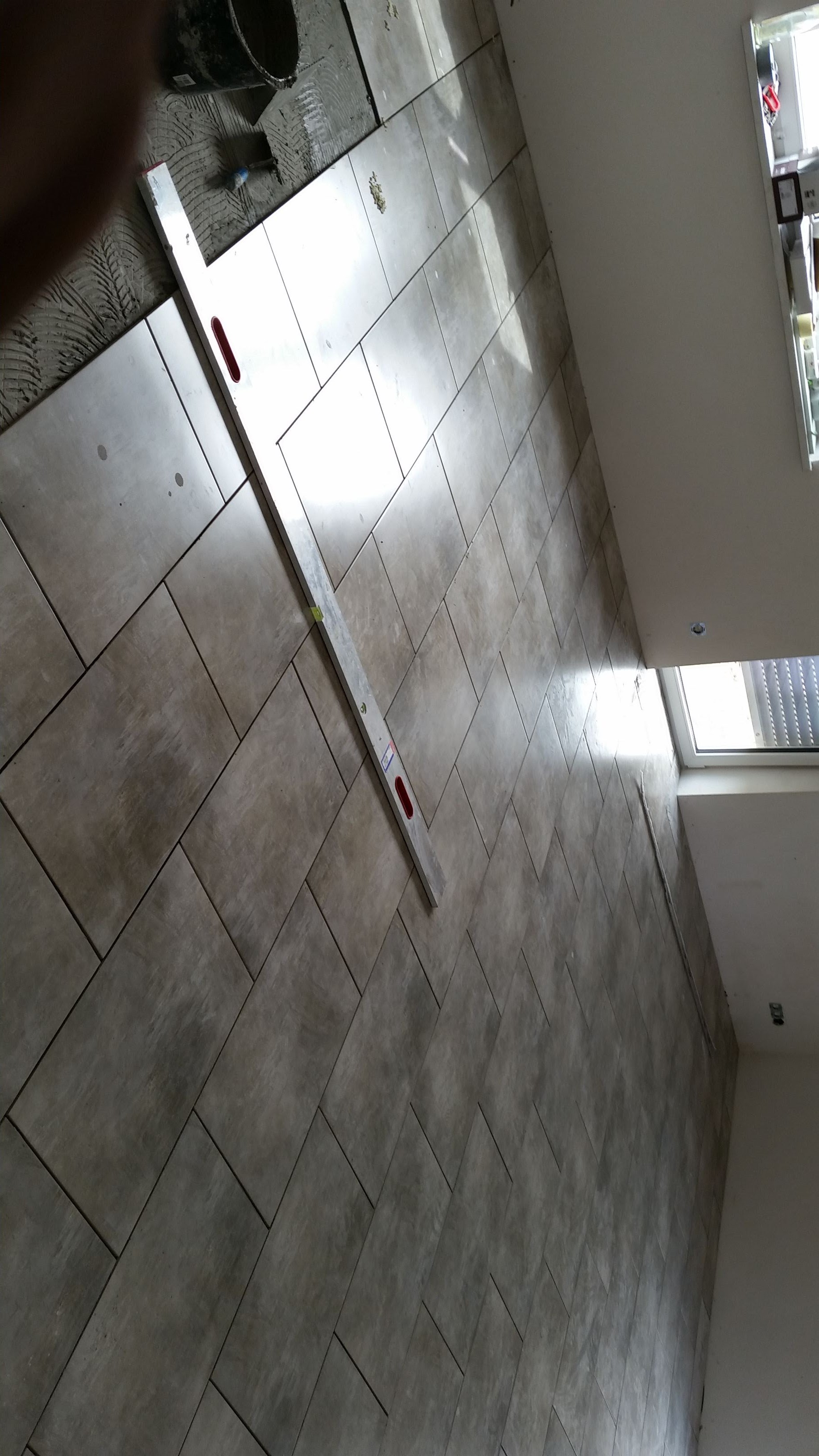 Układanie płytek podłogowych w romby, widoczna poziomica i wiadro z zaprawą, fragment okna w tle.