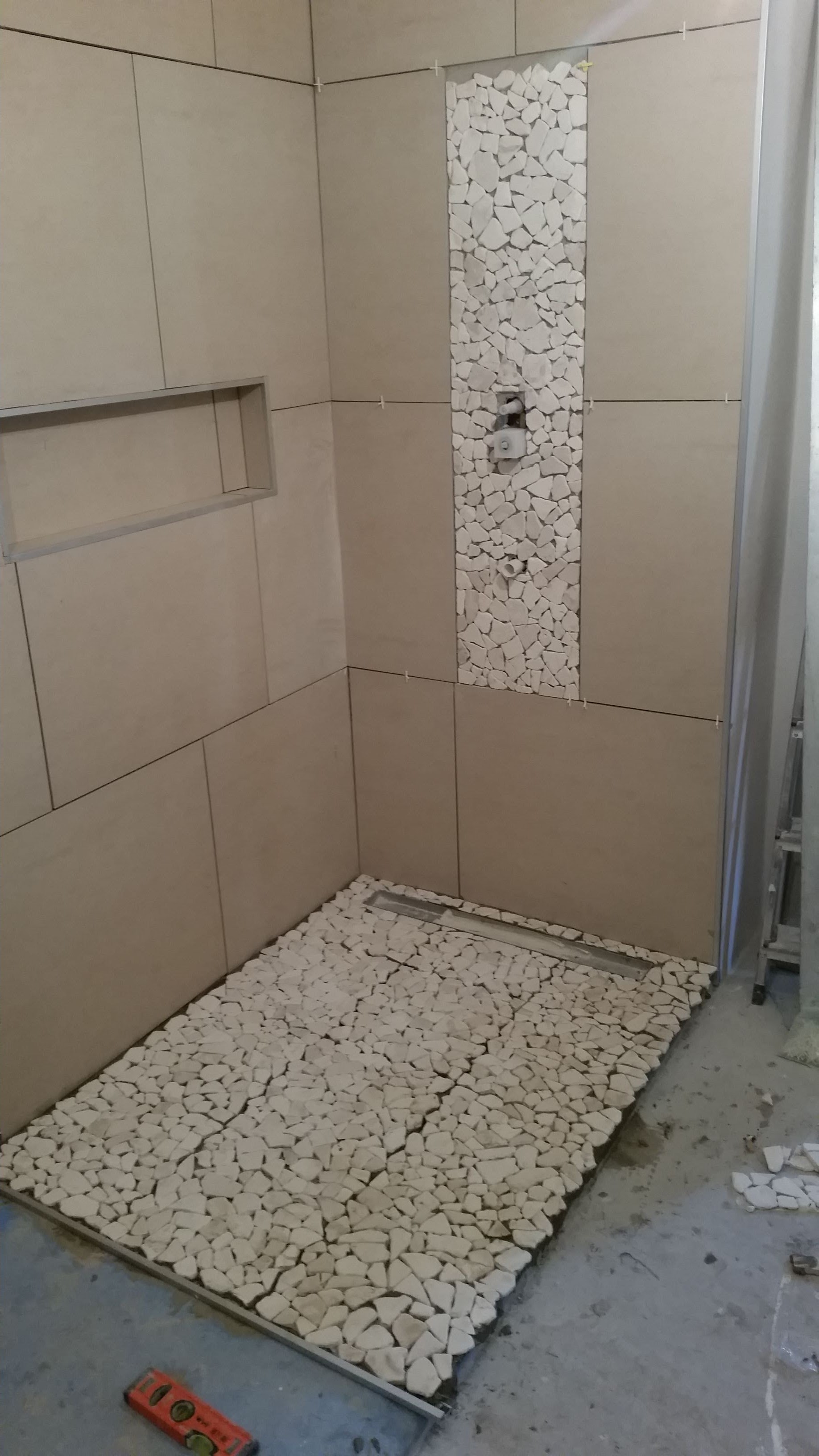 Niedokończona kabina prysznicowa z beżowymi płytkami ściennymi i mozaiką z białych kamyków na podłodze i pionowym pasie na ścianie; widoczna poziomica na podłodze.