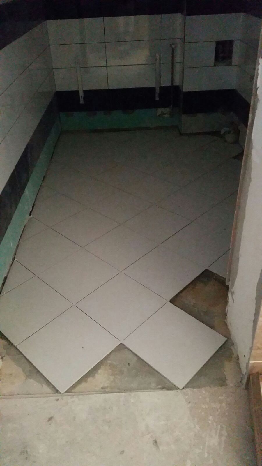 Układanie jasnych, kwadratowych płytek podłogowych w karo w pomieszczeniu z częściowo wykończonymi ścianami, widoczne niedokończone fragmenty podłogi i ściany z instalacjami.