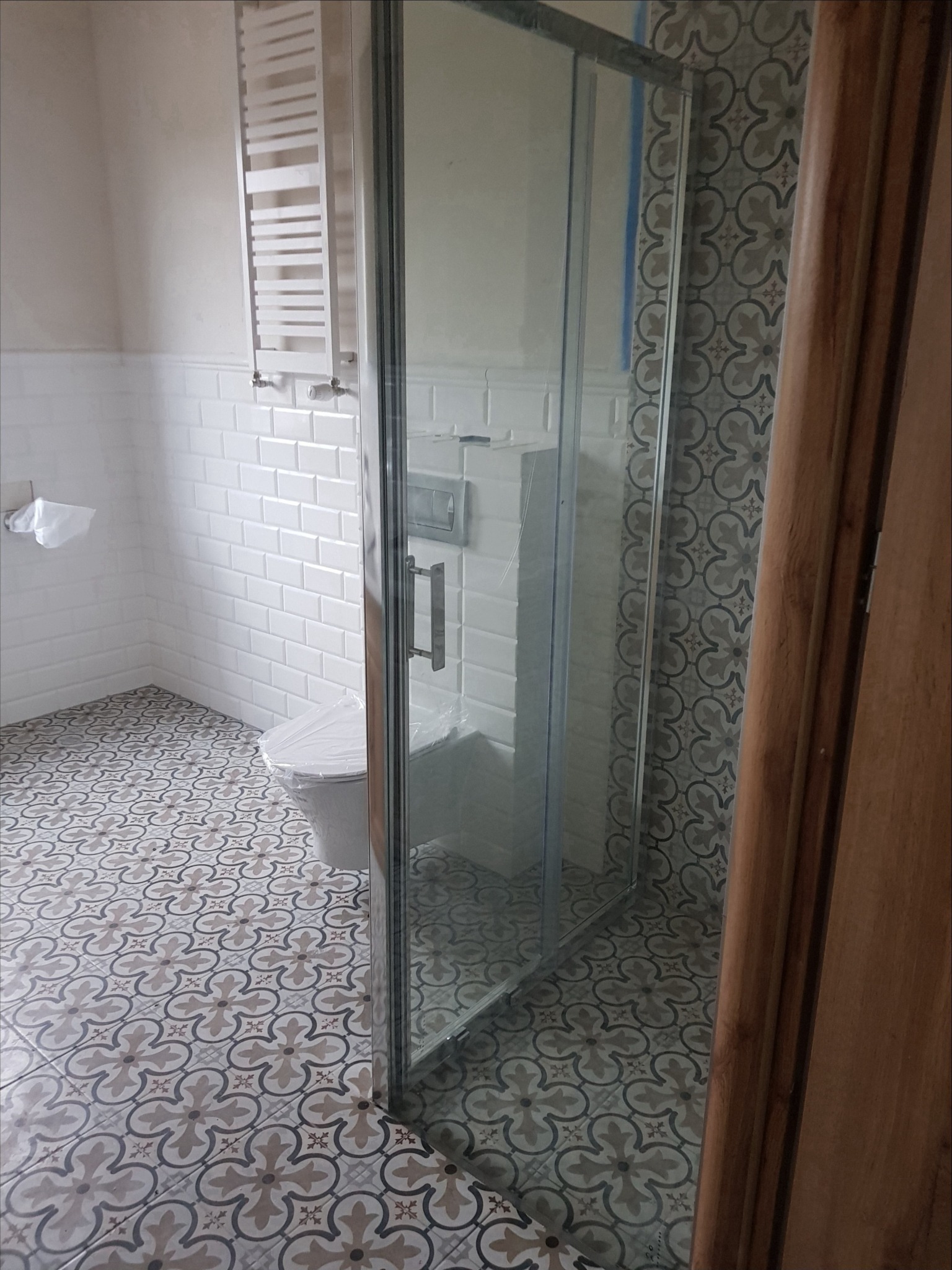 Ukończona łazienka z białą armaturą, kabiną prysznicową z przezroczystego szkła, grzejnikiem drabinkowym i wzorzystą podłogą w odcieniach szarości i beżu.