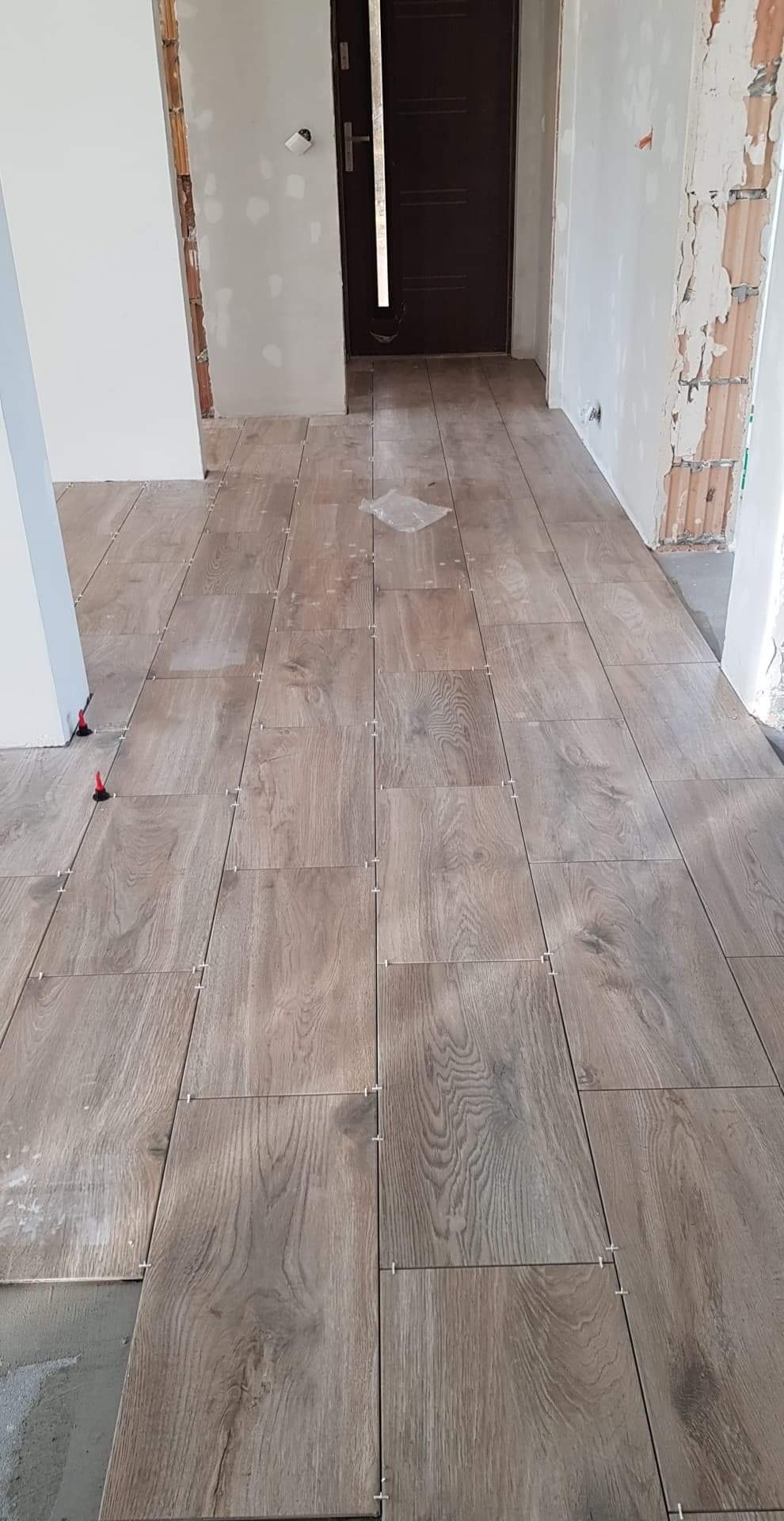 Układanie podłogi z płytek imitujących drewno, widoczne krzyżyki dystansowe i czerwone kliny poziomujące, surowe ściany w tle.