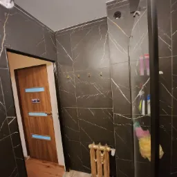 Łazienka z czarnymi płytkami imitującymi marmur ze złotymi żyłami, drewnianą podłogą i grzejnikiem w stylu retro, widok na drzwi z szybkami i kabinę prysznicową z czarną ramą.