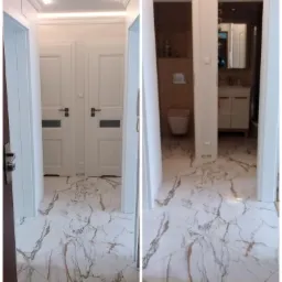 Korytarz z białymi drzwiami z przeszkleniami i marmurową podłogą w odcieniach bieli i złota, widok na wykończoną łazienkę z umywalką i toaletą.