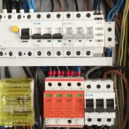 Rozdzielnica elektryczna z widocznymi bezpiecznikami Eaton, ogranicznikiem przepięć VCX w kolorze pomarańczowym oraz żółtym modułem LBR-100A/7, okablowanie w różnych kolorach.