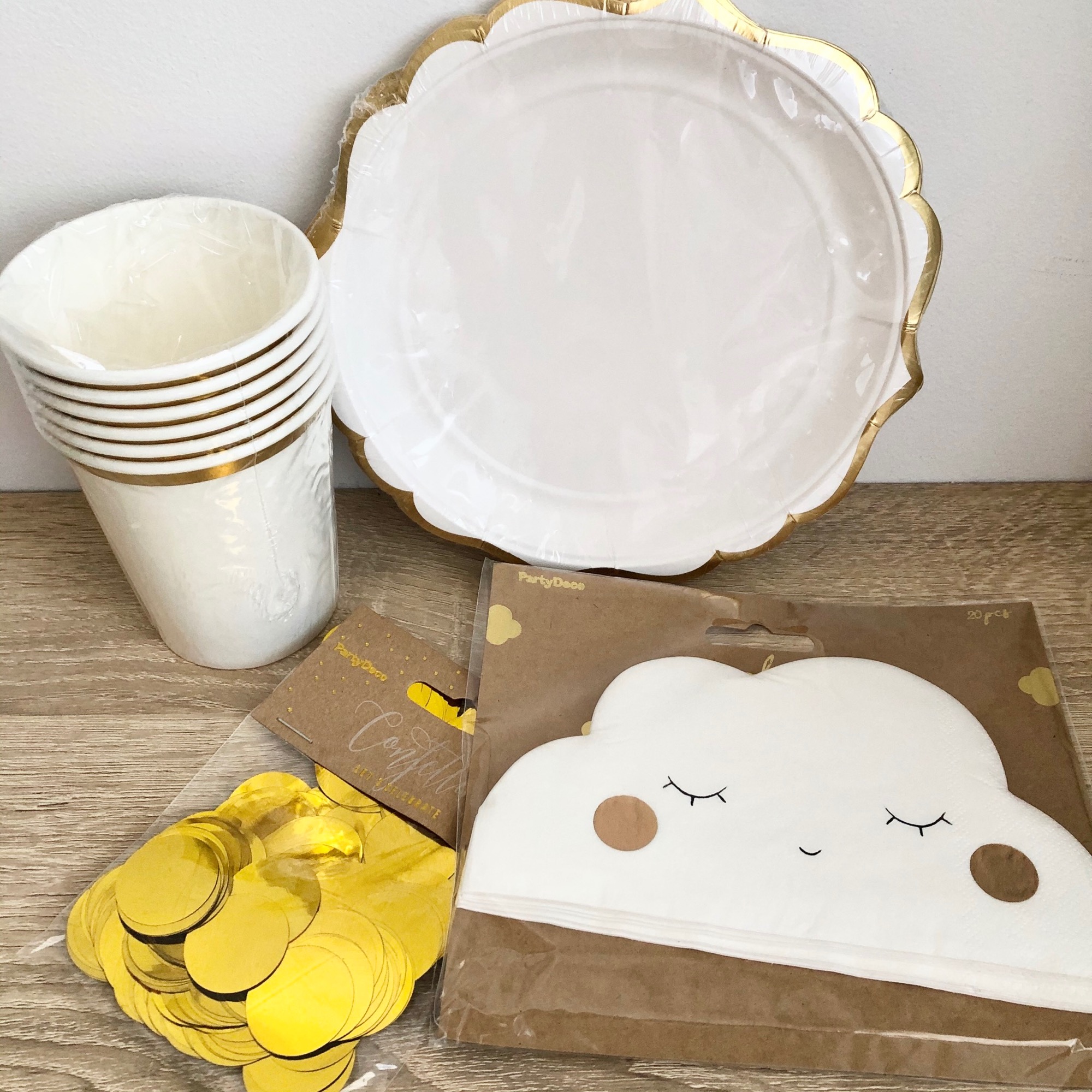 Zestaw imprezowy: białe talerze z falowaną złotą obwódką, papierowe kubeczki z złotymi paskami, konfetti w złotym kolorze oraz serwetki w kształcie chmurek z zamkniętymi oczami, zapakowane w folię.