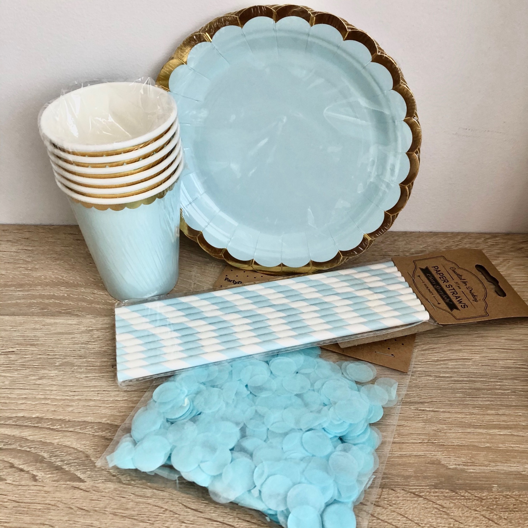 Zestaw imprezowy: błękitne papierowe talerzyki ze złotym brzegiem, kubeczki, słomki w paski i konfetti w przezroczystym opakowaniu, ułożone na drewnianej powierzchni.