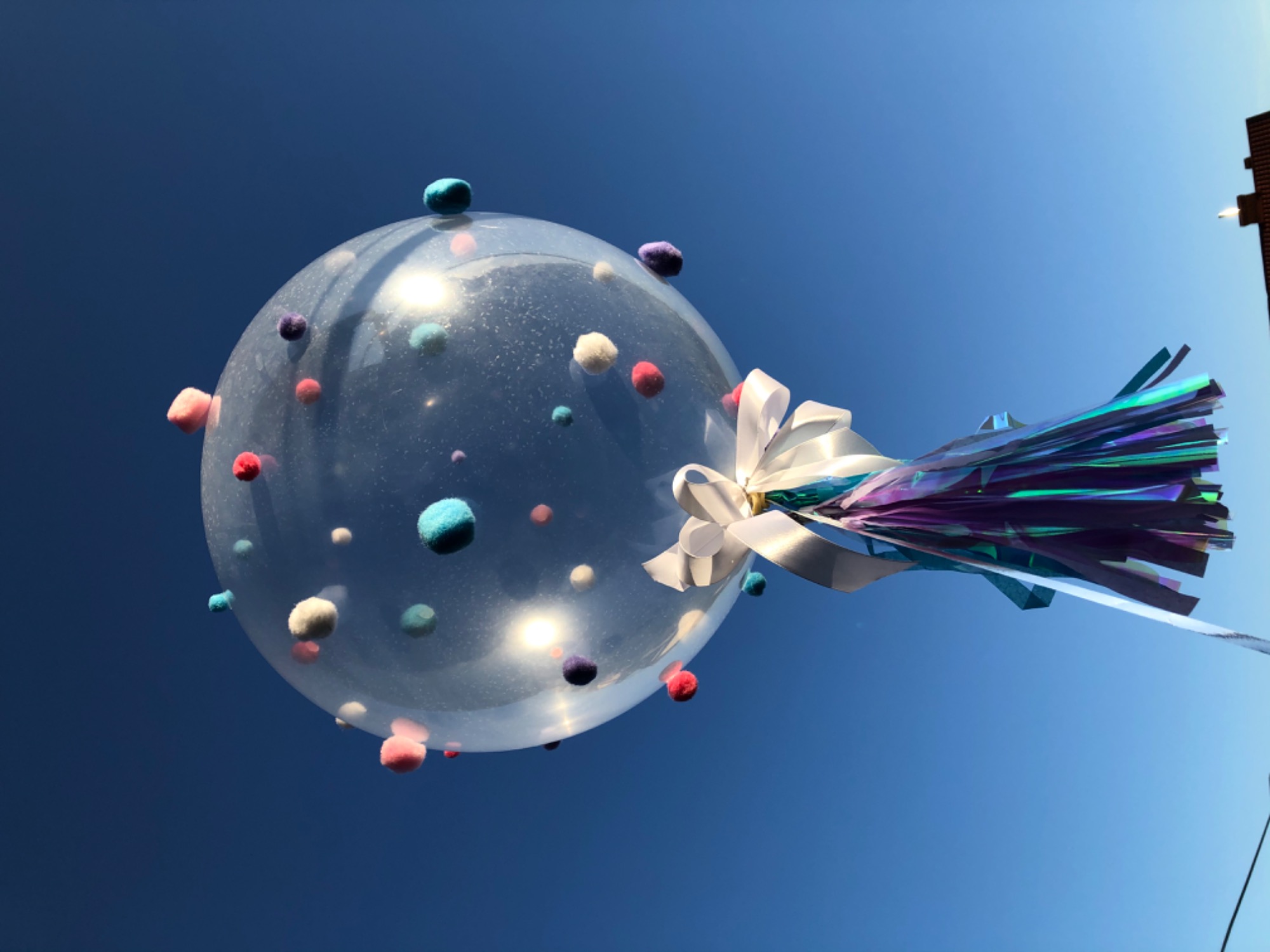 Przezroczysty balon wypełniony kolorowymi pomponami, zawiązany białą kokardą i holograficznymi frędzlami, na tle błękitnego nieba.