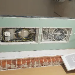 Dwa otwory w płycie gipsowej ukazujące instalacje elektryczne w trakcie montażu, z widocznymi przewodami, puszkami i elementami montażowymi, surowa ceglana ściana w tle.