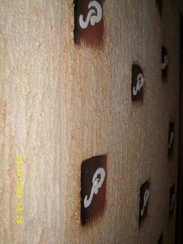 Teksturowana ściana z tynkiem akrylowym typu kornik w odcieniach beżu, ozdobiona ciemnobrązowymi kwadratami z białymi, stylizowanymi literami 'S'.