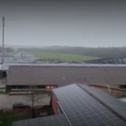 Widok z góry na budynki gospodarcze z zainstalowanymi panelami słonecznymi na dachach, w tle pola uprawne i las, pochmurny dzień.