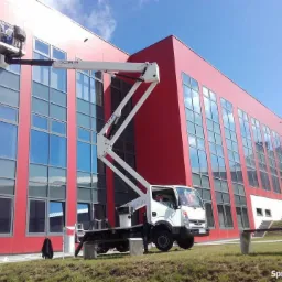 Pracownicy na podnośniku koszowym czyszczą elewację budynku z czerwonymi panelami i dużymi oknami.