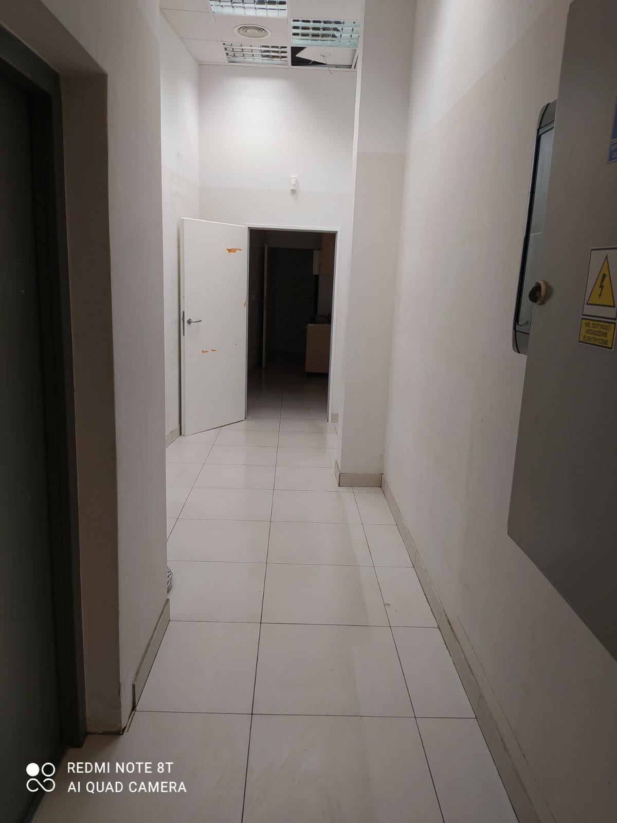 Wnętrze korytarza z białymi płytkami podłogowymi, otwartymi drzwiami w tle i szarą szafką elektryczną po prawej stronie z żółtą naklejką ostrzegawczą.