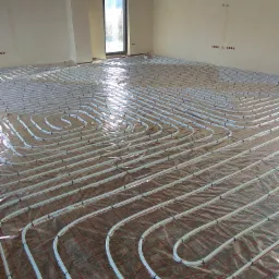 Białe rury ogrzewania podłogowego ułożone w pętle na folii z czerwoną kratką w surowym wnętrzu z oknem.