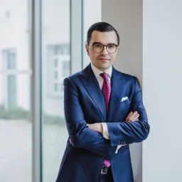 Portret mężczyzny w okularach i granatowym garniturze z fioletowym krawatem w kropki, opierającego się o białą ścianę na tle okna.