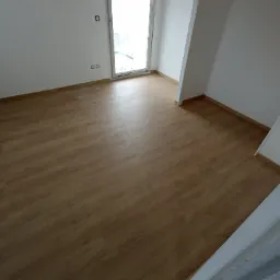 Jasne panele podłogowe w pustym, nowo wykończonym pokoju z białymi ścianami, widoczne gniazdka elektryczne i otwarte okno balkonowe.