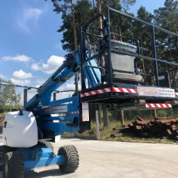 Niebiesko-biały podnośnik koszowy na betonowej powierzchni, z widocznym koszem roboczym, oponami terenowymi i lasem w tle, pod błękitnym niebem z lekkimi chmurami.
