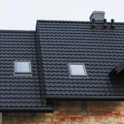 Czarny dach z dachówki metalowej z dwoma oknami dachowymi i kominem na budynku w stanie surowym, z widocznymi cegłami.