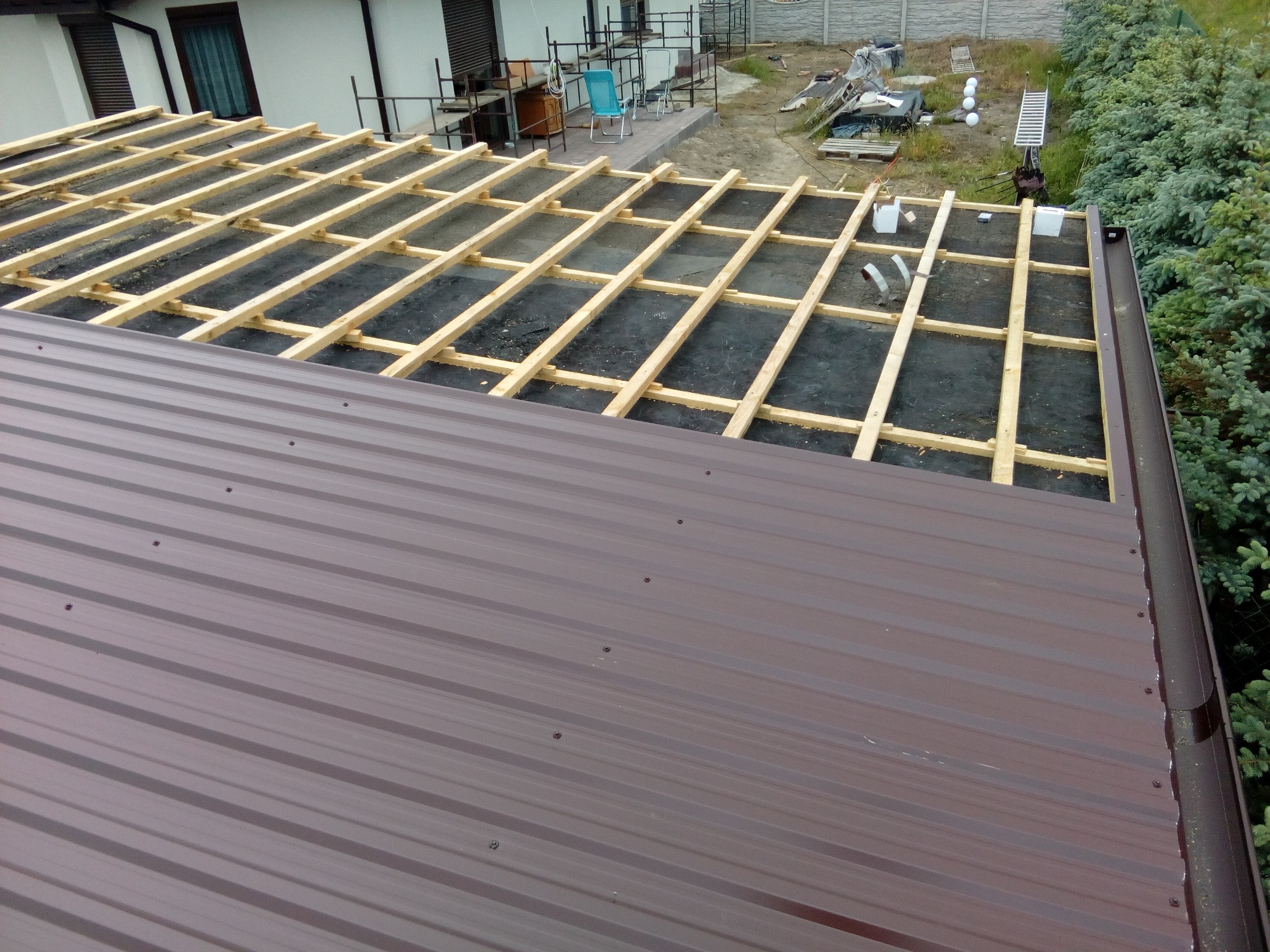 Częściowo pokryty brązową blachą dach z widoczną drewnianą konstrukcją łat i kontrłat na czarnej membranie dachowej. W tle dom z szarymi okiennicami i ogród.