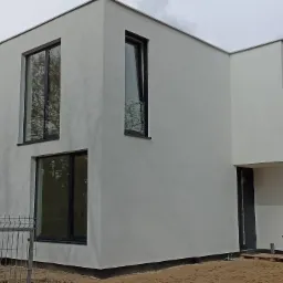 Nowoczesny, biały dom z czarnymi ramami okiennymi, widok z zewnątrz, częściowo ogrodzony, w tle drzewa i pochmurne niebo.