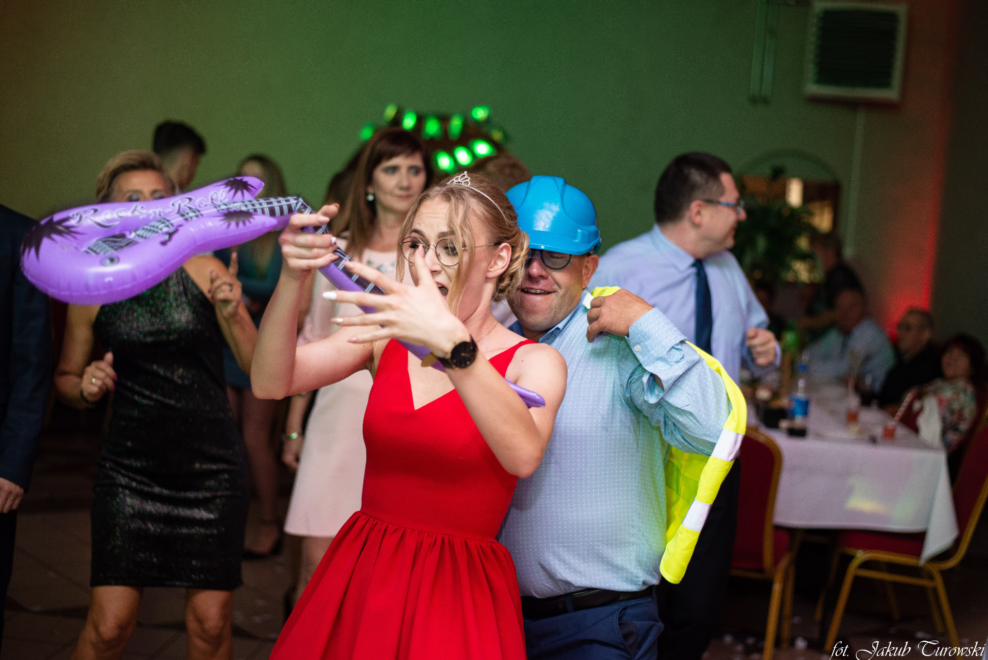 Rozbawiona kobieta w czerwonej sukience i tiarze pozuje z dmuchaną gitarą na imprezie, obok mężczyzna w kasku budowlanym i kamizelce odblaskowej.