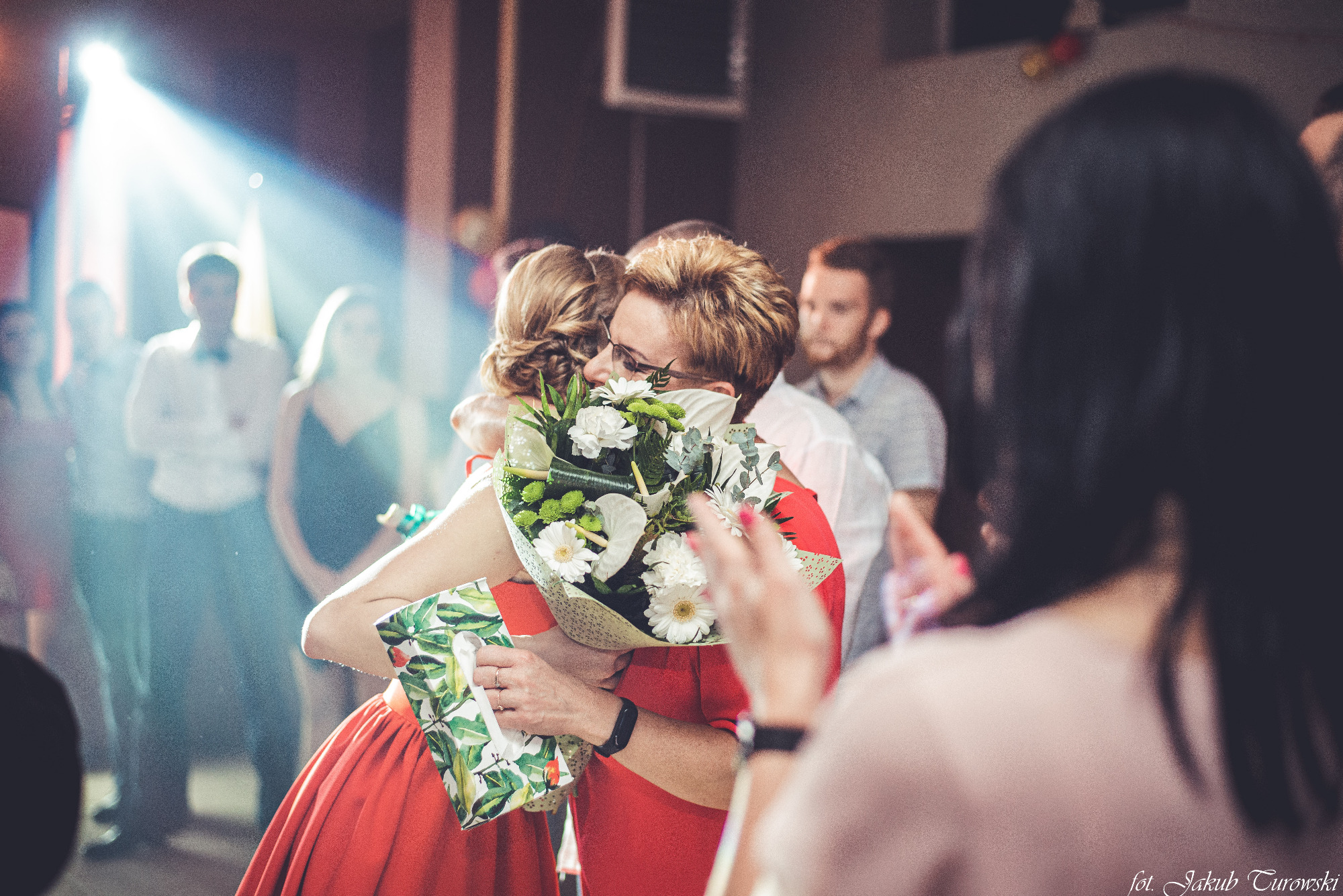 Dwie kobiety w czerwonych sukienkach obejmują się trzymając bukiet białych kwiatów i chusteczki higieniczne na tle rozmytych gości podczas wydarzenia.