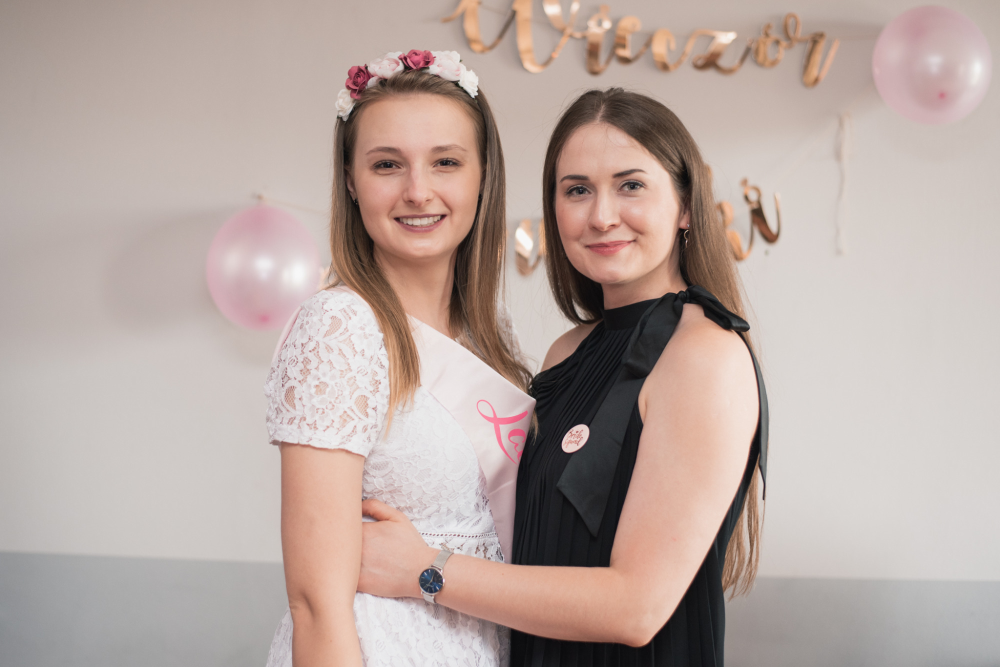 Dwie uśmiechnięte kobiety, jedna w koronkowej sukience i z wiankiem, druga w czarnej sukience, pozują na tle dekoracji urodzinowych z balonami i złotymi napisami.