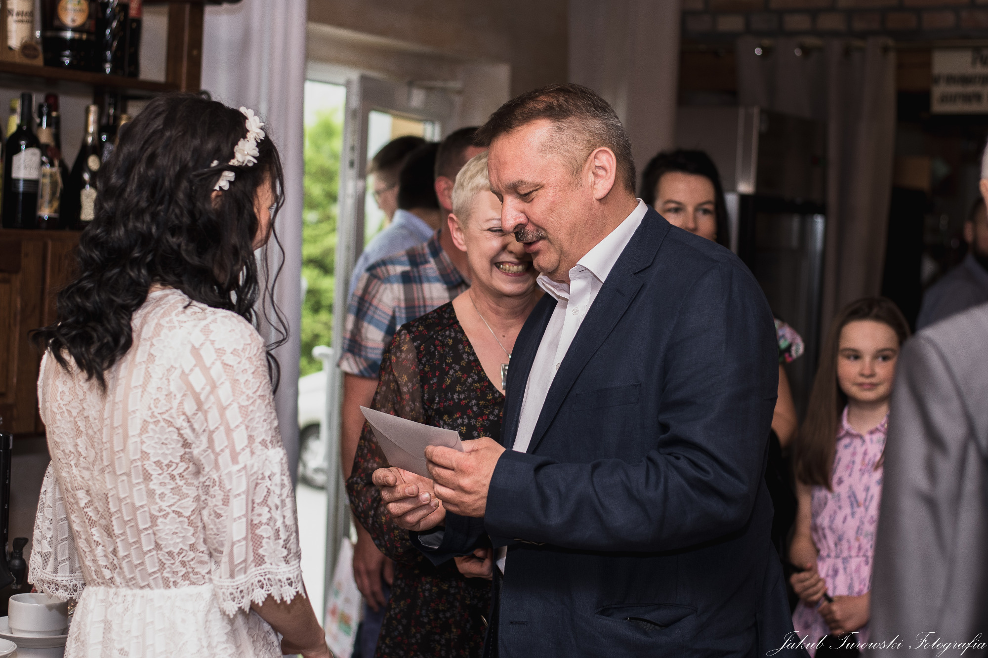 Goście weselni wręczają kopertę z prezentem pannie młodej w koronkowej sukni, widoczna uśmiechnięta kobieta i dziewczynka w tle.