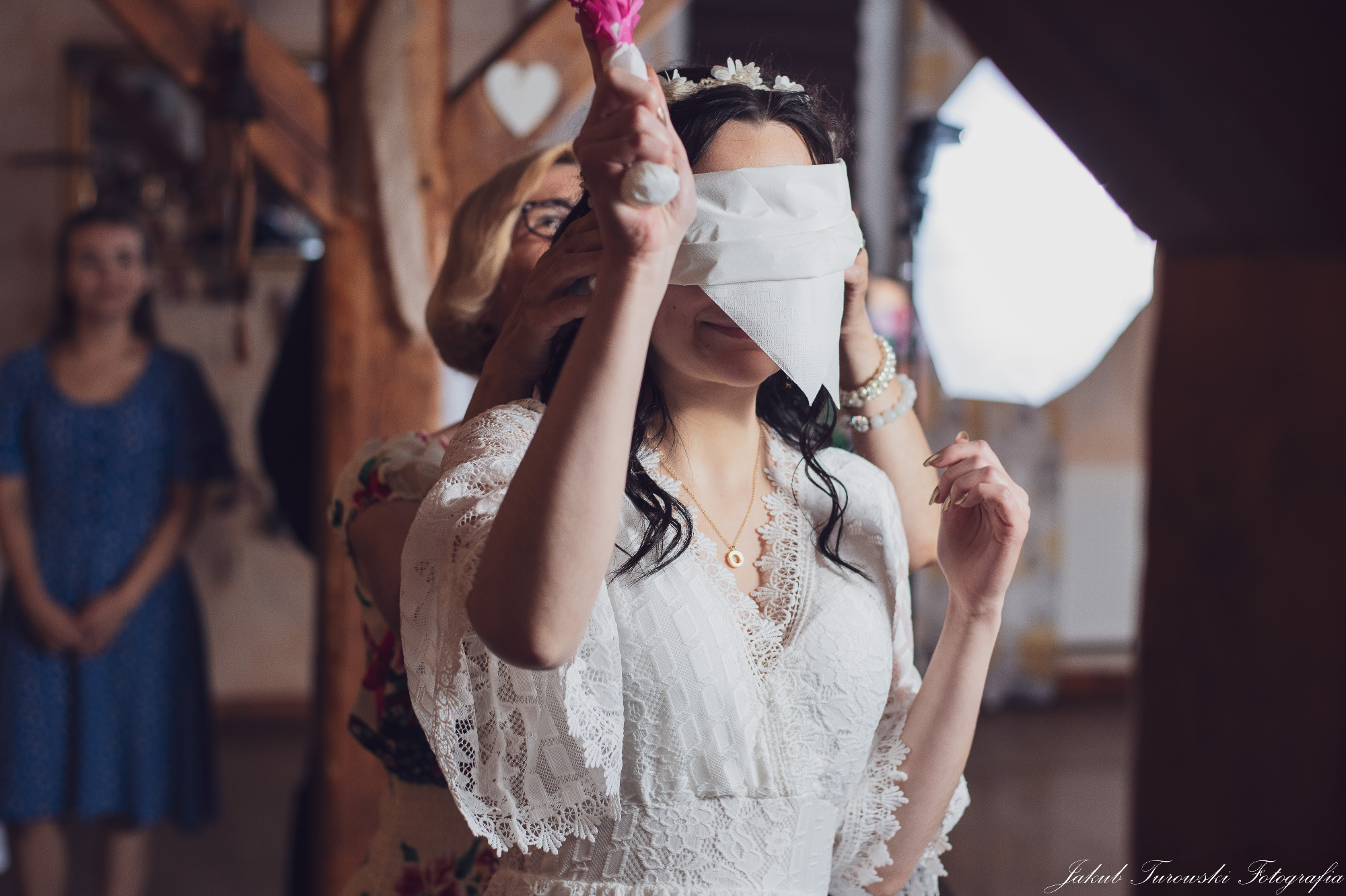 Panna młoda z zawiązanymi oczami podczas oczepin, trzymana przez kobietę w kwiecistej sukience, w tle widoczna inna kobieta i drewniana konstrukcja.