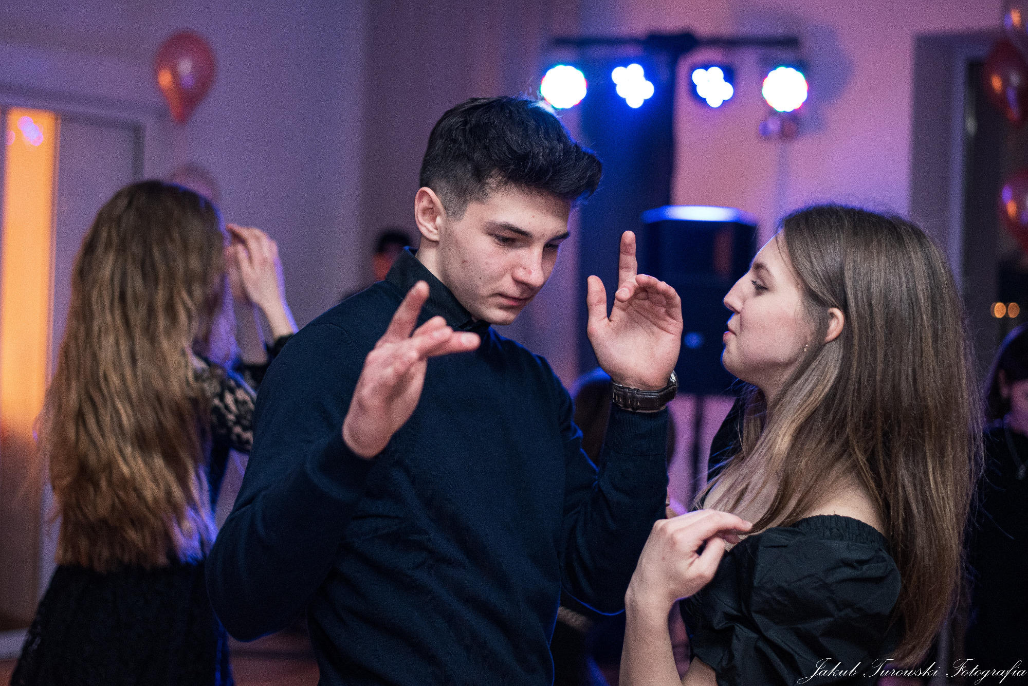 Młody mężczyzna w granatowym swetrze gestykuluje rękami podczas wydarzenia, w tle tańcząca kobieta i efekty świetlne.