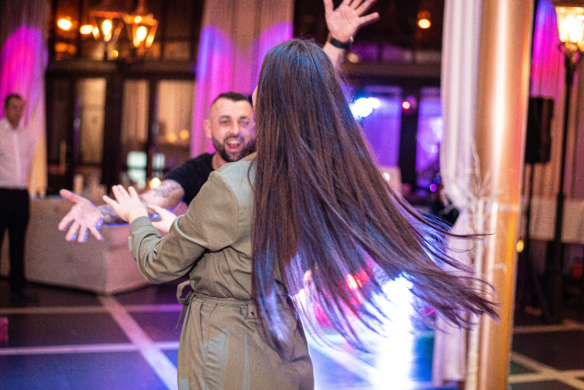 Rozmazane ujęcie tańczącej pary na evencie, z długimi, ciemnymi włosami kobiety w ruchu, oświetlonymi niebieskim światłem, oraz uśmiechniętym mężczyzną z tatuażami w tle.