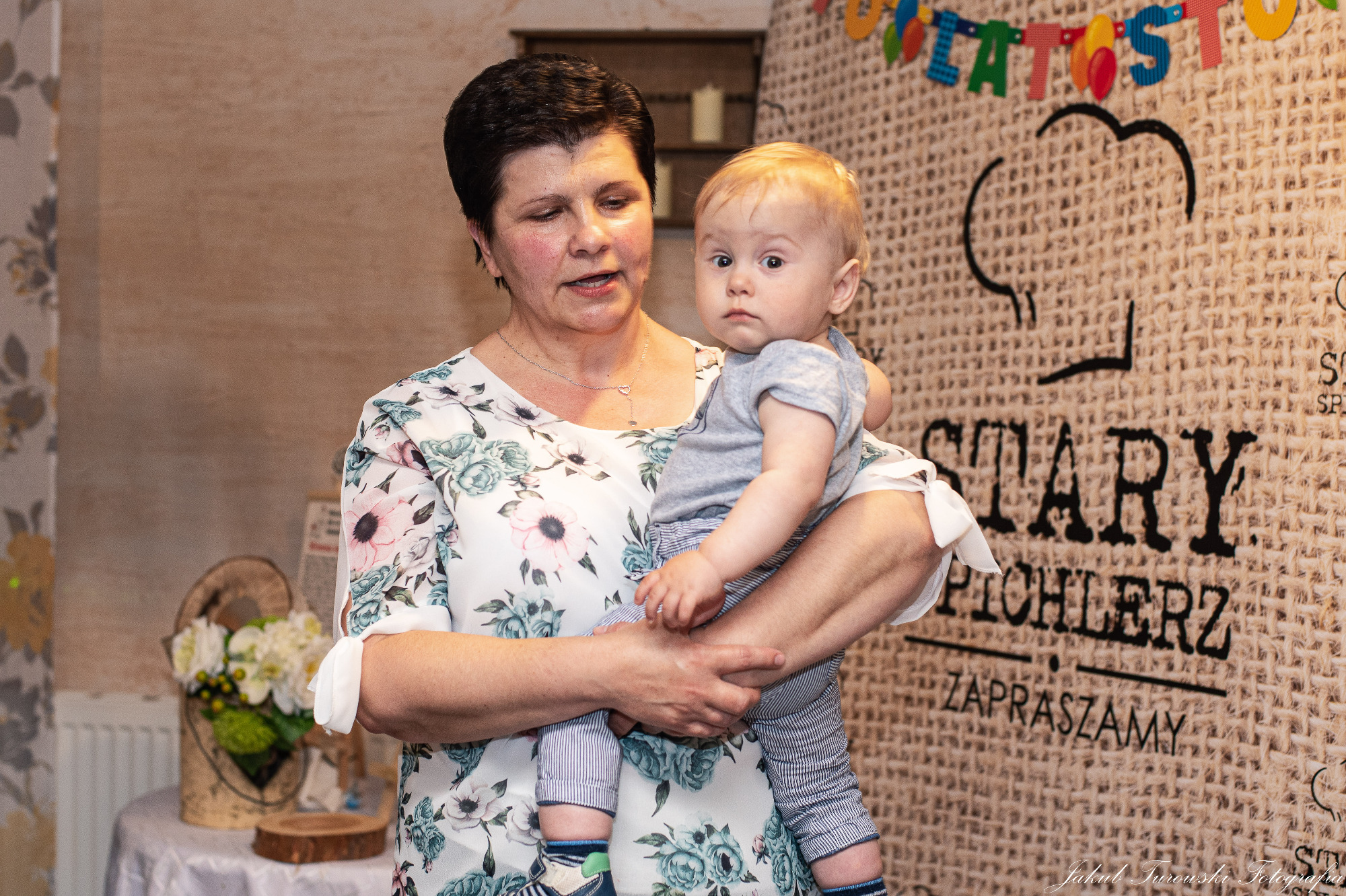Kobieta w kwiecistej bluzce trzyma na rękach małego chłopca w paski na tle ściany z napisem 'Stary Pichlerz Zapraszamy' i dekoracją urodzinową.