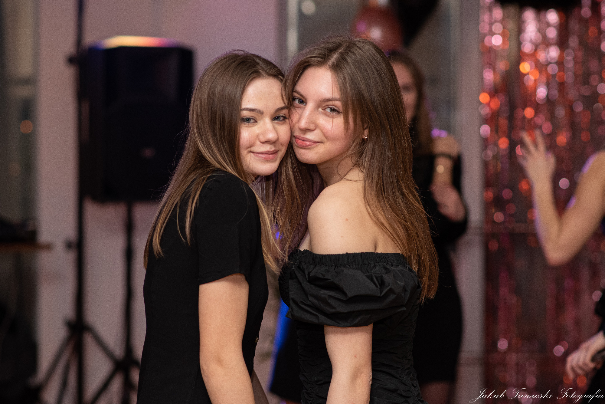 Dwie uśmiechnięte nastolatki w czarnych sukienkach pozują blisko siebie na tle dekoracji imprezowej z błyszczących frędzli.