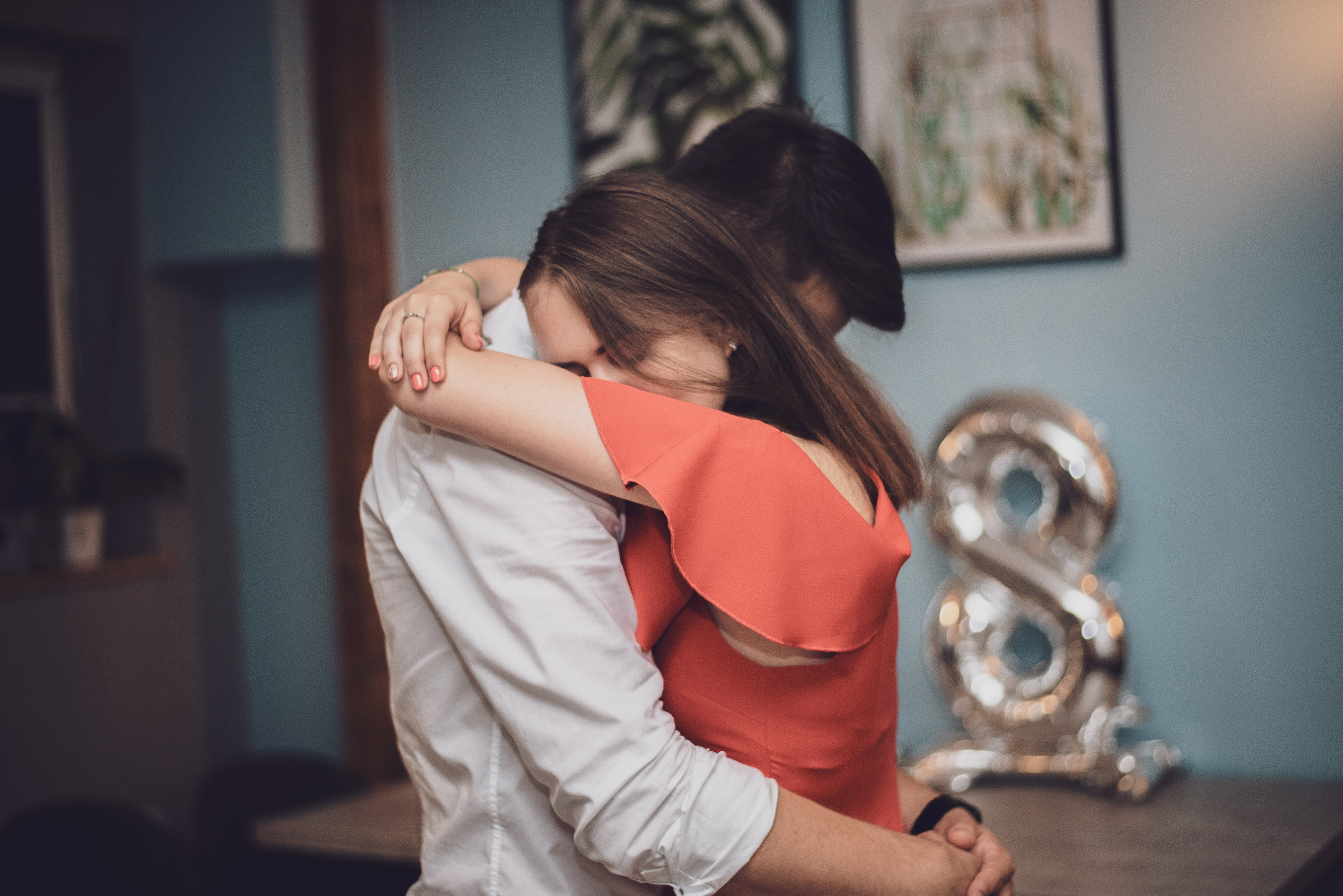 Para obejmuje się w pomieszczeniu z balonem w kształcie cyfry osiem, kobieta w czerwonej sukience z odkrytymi ramionami, mężczyzna w białej koszuli.