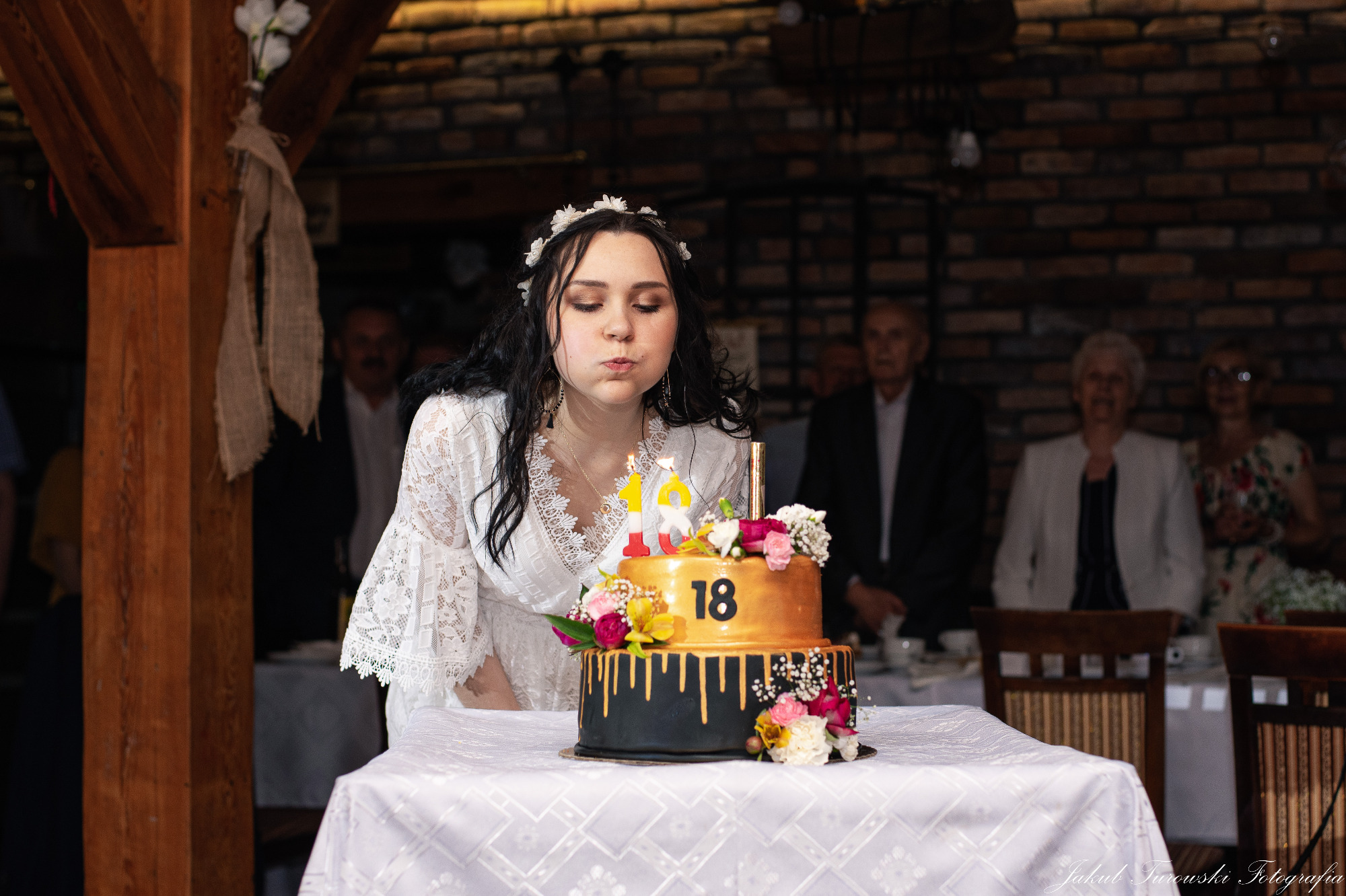 Młoda kobieta w białej koronkowej sukience, z wiankiem na głowie, zdmuchuje świeczki z tortu urodzinowego z numerem 18, na tle ceglanej ściany i osób siedzących przy stole.