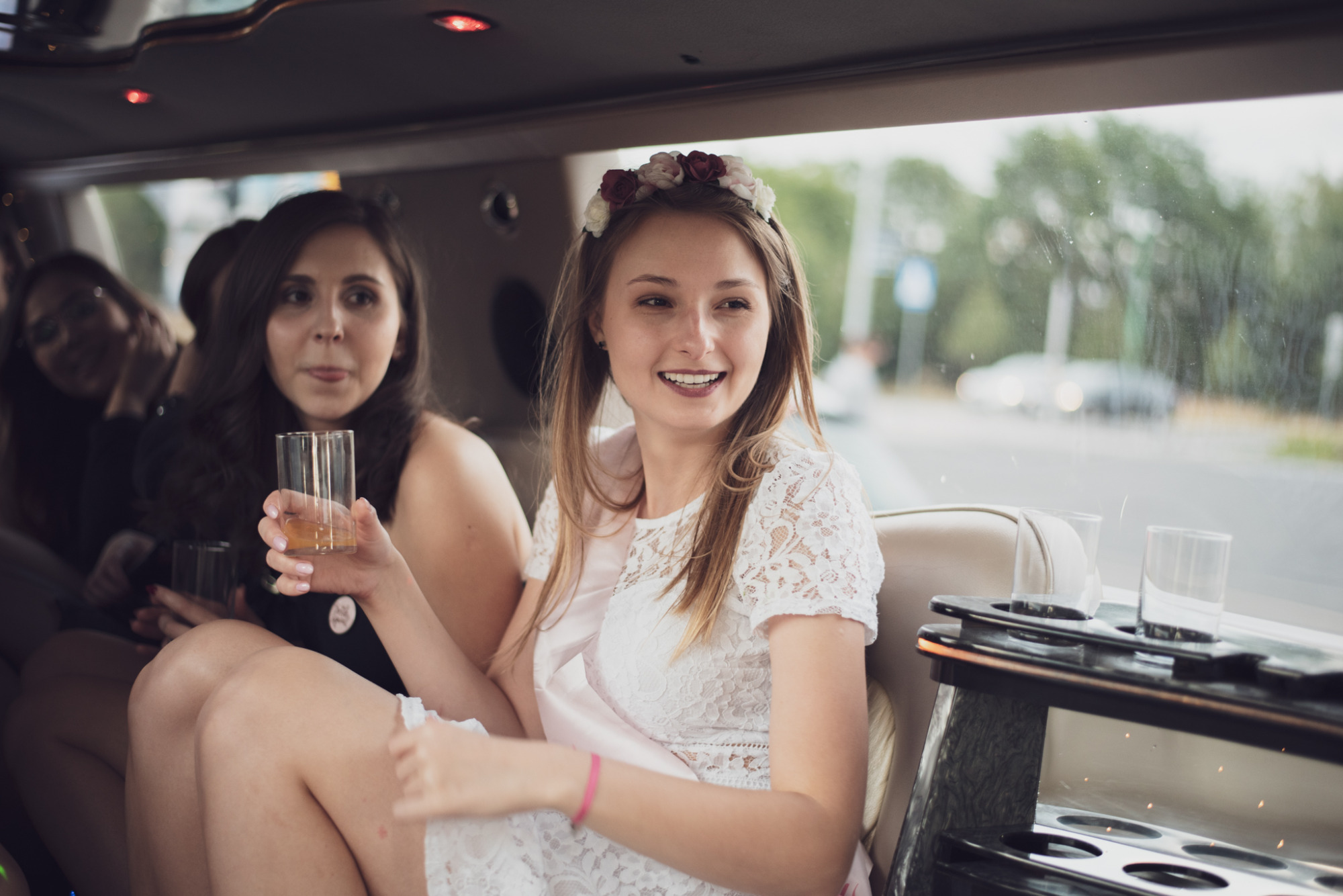 Uśmiechnięta panna młoda w koronie z kwiatów i białej koronkowej sukience, siedzi w limuzynie z przyjaciółkami, trzymając szklankę z napojem.