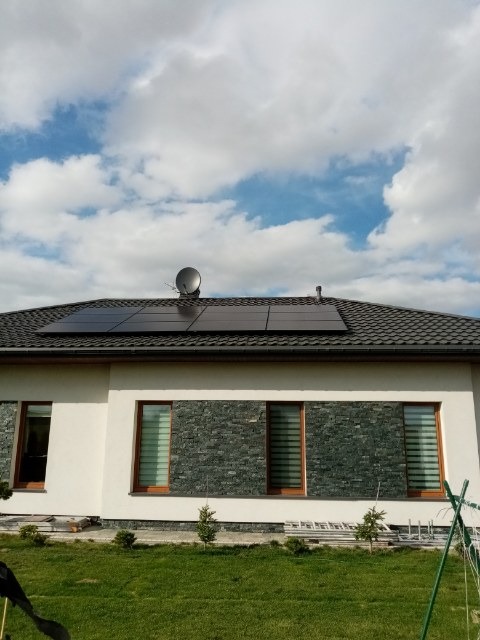 Panele słoneczne na dachu domu jednorodzinnego z anteną satelitarną, widok z ogrodu, częściowo zachmurzone niebo.