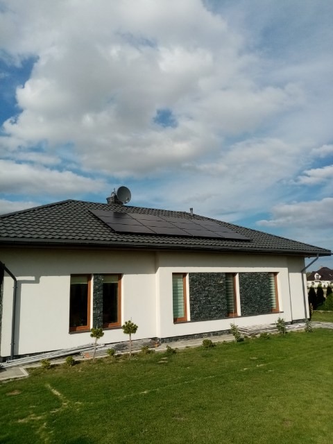 Dom jednorodzinny z zainstalowanymi panelami fotowoltaicznymi na ciemnym dachu, widoczne również antena satelitarna, biała elewacja, zielony trawnik i dekoracyjne drzewka iglaste.