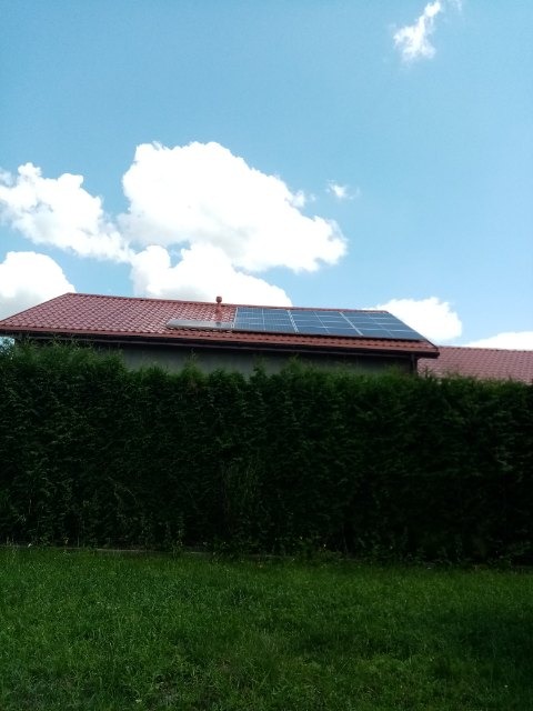 Panel fotowoltaiczny zamontowany na dachu domu z czerwoną dachówką, widok z poziomu trawnika z żywopłotem w tle, błękitne niebo z białymi chmurami.